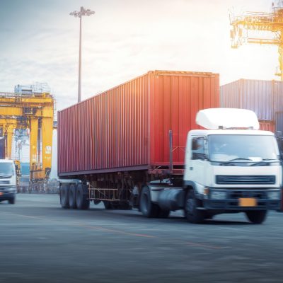 A white truck with a red container drives through a shipping port with other trucks, cranes, and stacks of containers in the background.